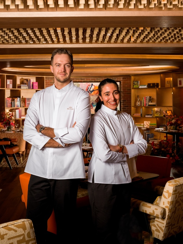 Two chefs in white uniforms pose confidently against a mural backdrop in a restaurant setting.