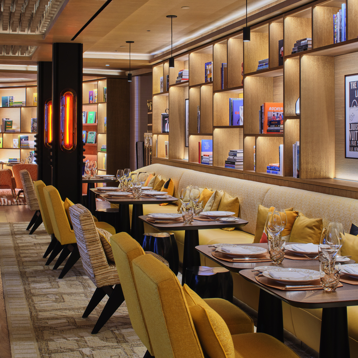 Modern restaurant interior with wooden floors, patterned chairs, orange accents, and a bar in the background.